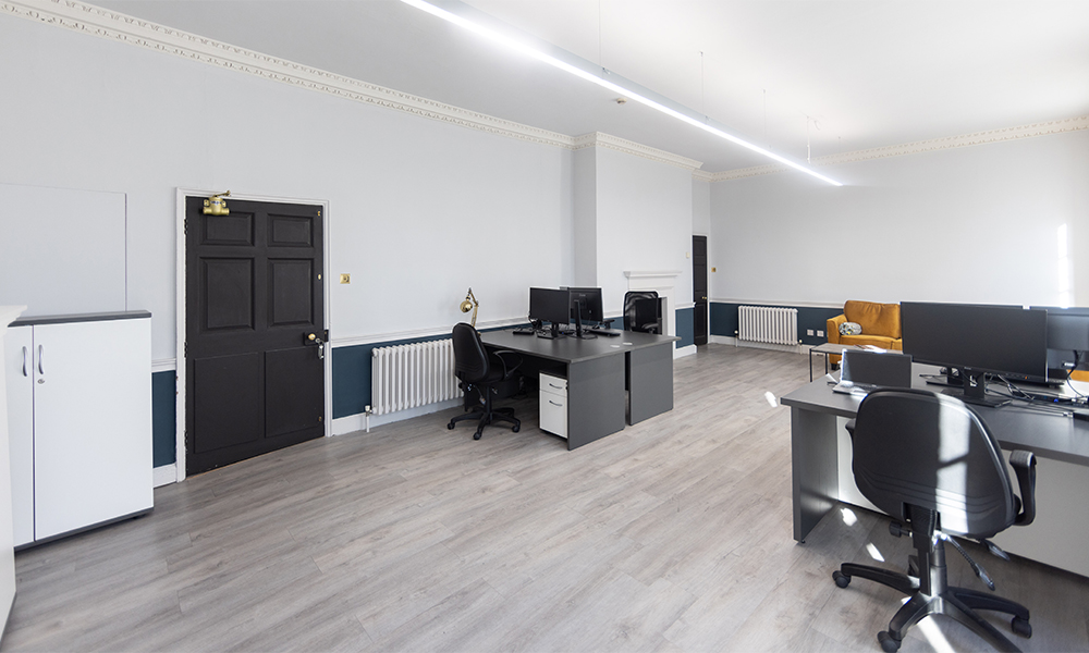 Spacious office with sleek desks, computers, and black chairs on light wood floors. A mustard sofa sits in the back corner, with bright lighting and a modern, minimalistic feel.